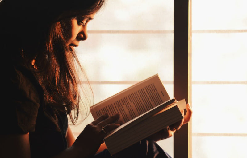 A woman engrossed in reading a book by a sunlit window creates a serene atmosphere.