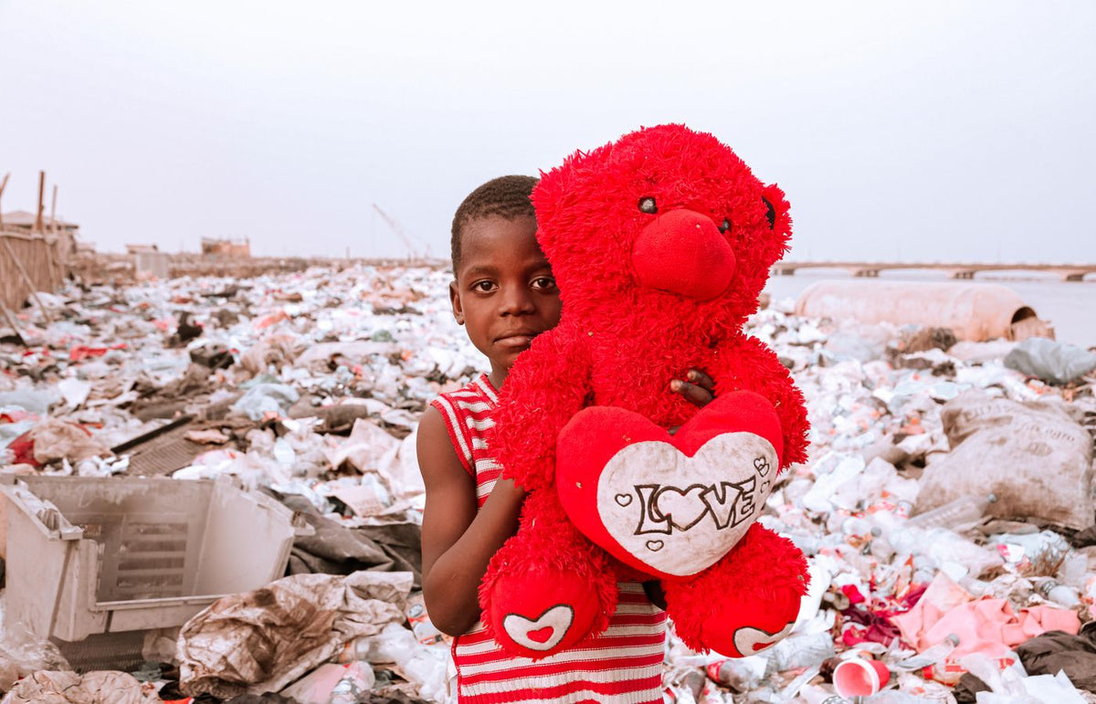 Young boy in a striped shirt holding a teddy bear amidst a landfill, highlighting waste issues.