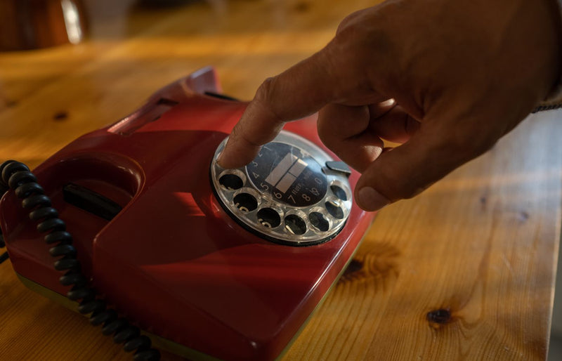 Close-up of a hand dialing a red rotary phone on a wooden table.