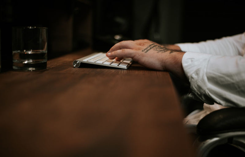 A close-up of a man's tattooed hands typing on a keyboard at a wooden desk.