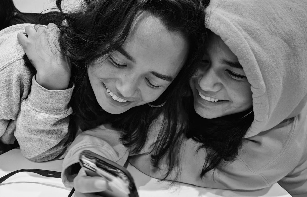 Two teenage girls laughing and smiling while looking at a smartphone in a black and white photo.
