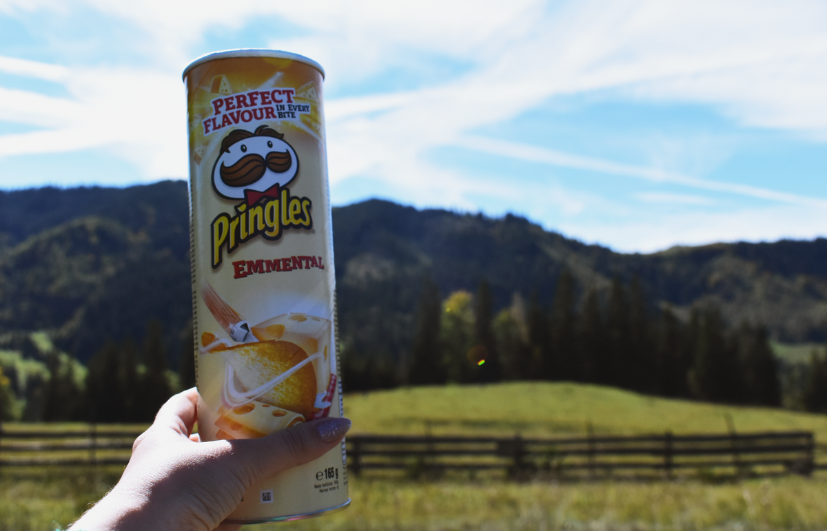 A hand holds a Pringles can against a scenic mountain backdrop with clear skies.