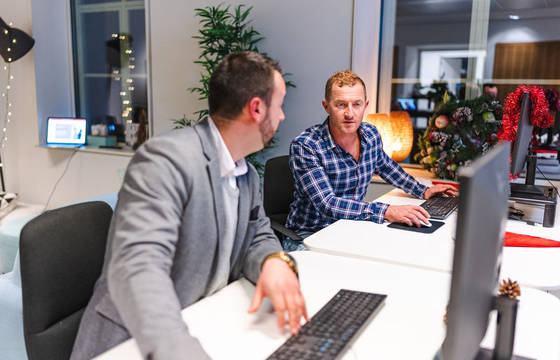 Two men collaborate in a modern office with computers and holiday decor.