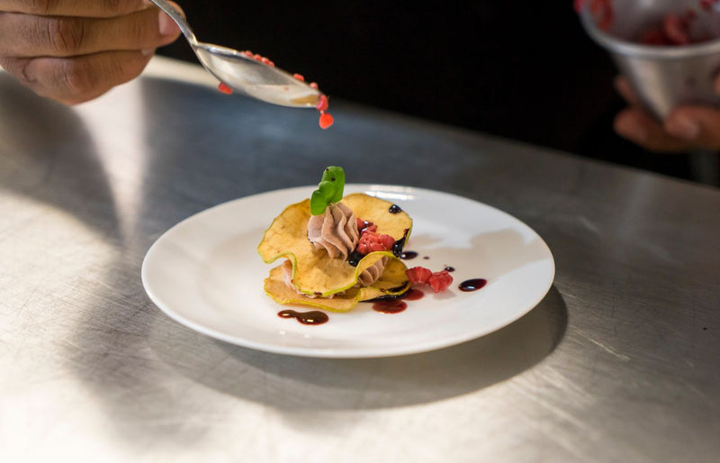 Close-up of a chef garnishing an elegant dessert with berry sauce on a white plate.