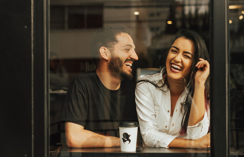 A man and woman laughing together at a café window with coffee.