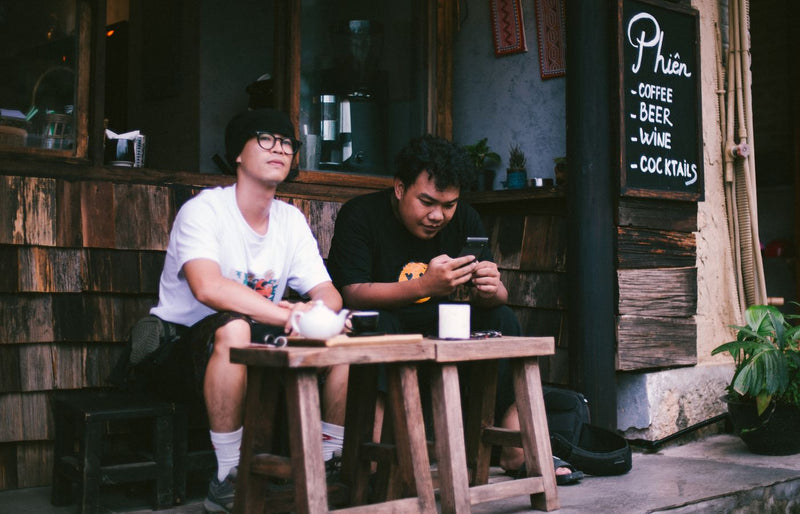 Two men sitting outside a café, enjoying coffee and using smartphones.