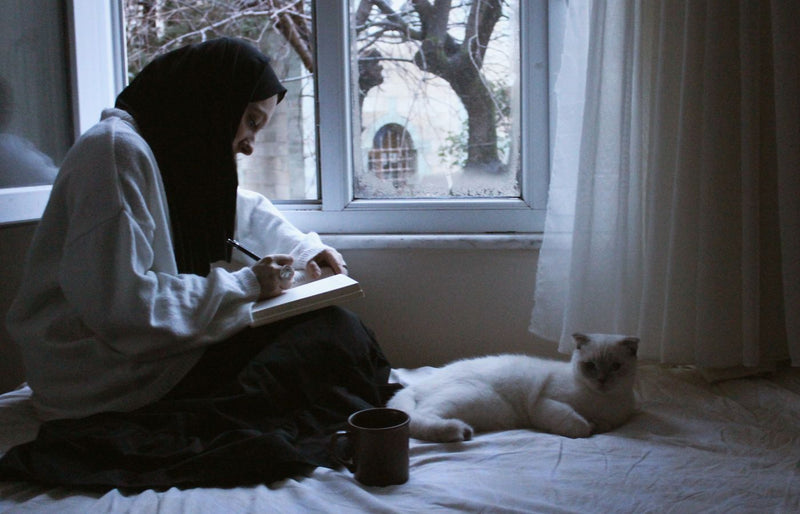 Muslim woman writing in a notebook next to a white cat, sitting by a window.