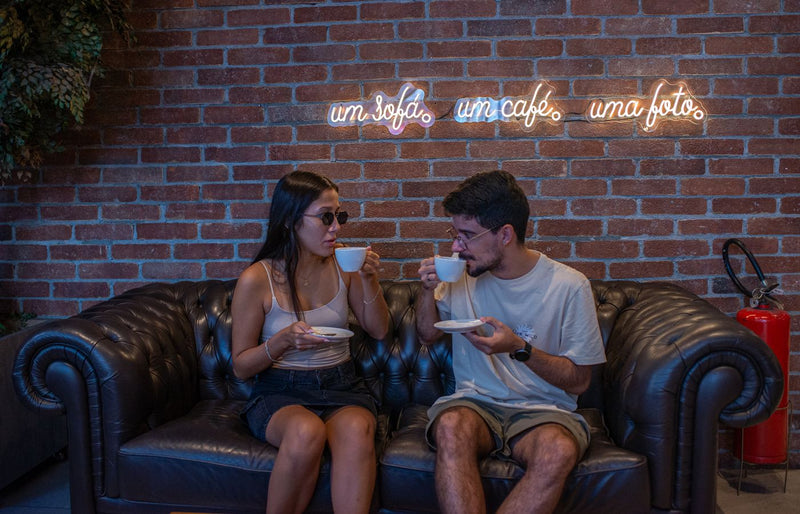 A couple enjoying coffee on a sofa in a trendy café with neon signs on a brick wall.