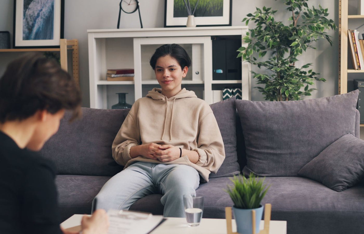 Teenager in therapy session sitting on a sofa, discussing with a professional therapist in a modern office setting.