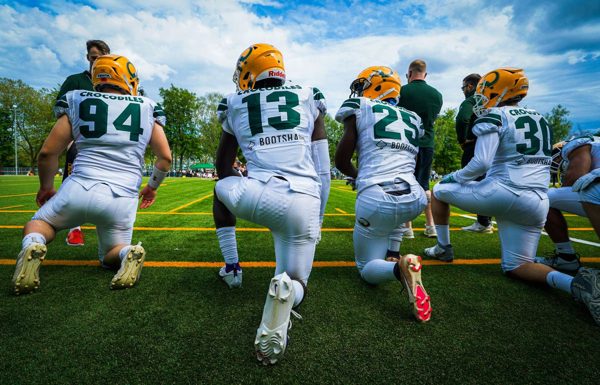 A group of American football players wearing helmets and sports gear kneeling on a green field.
