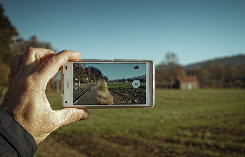 A hand holds a smartphone, capturing a clear image of a scenic countryside road and field.