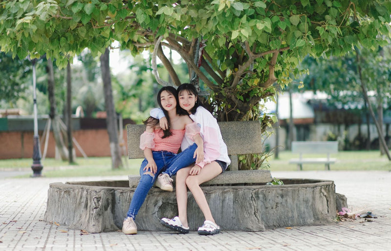 Two women sitting and hugging under a tree in a sunny park, exuding joy and friendship.