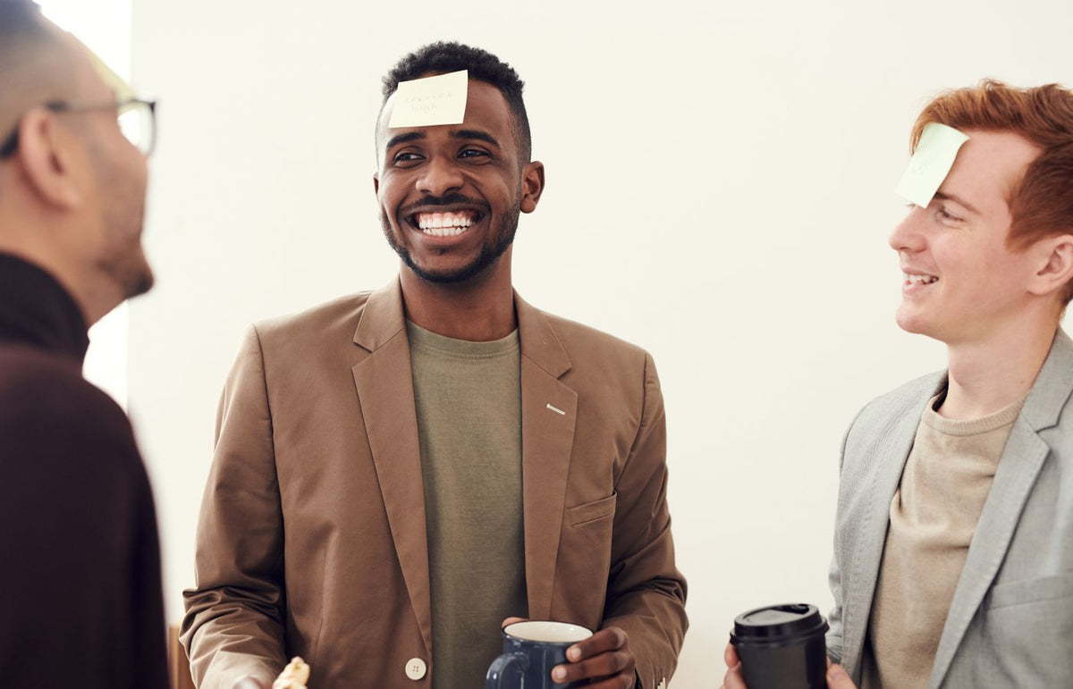 Three men laughing and playing a fun game with sticky notes indoors.