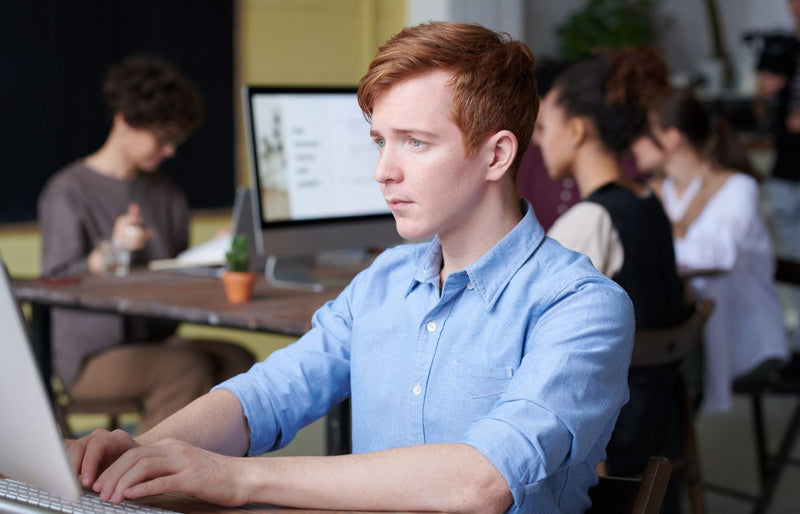 Young professional working intently on a computer in a busy modern office setting, surrounded by colleagues.