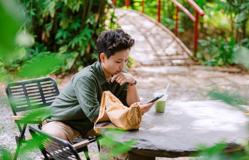 Woman sitting outdoors with a phone and a drink, enjoying a relaxing break in nature.
