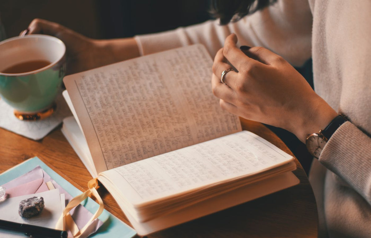 A woman writes in a journal while enjoying a cup of coffee at a wooden table.