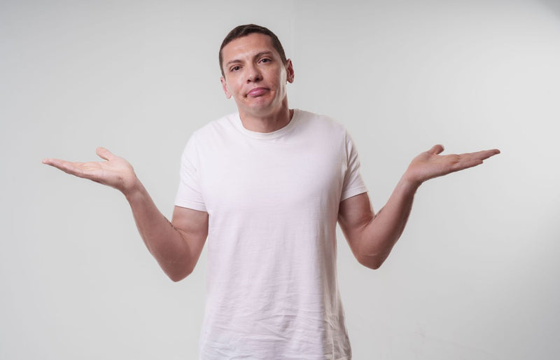 An adult man wearing a white t-shirt shrugs against a neutral background, conveying confusion or indifference.