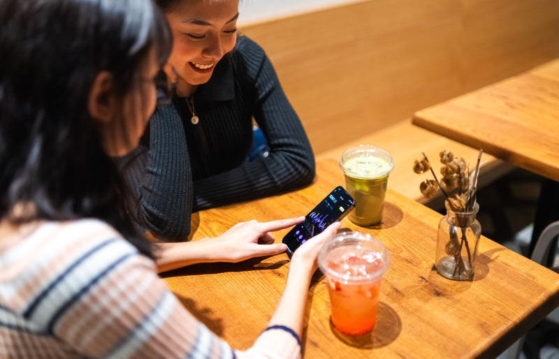 Two friends enjoy drinks at a cafe while looking at a stock market app on a smartphone.