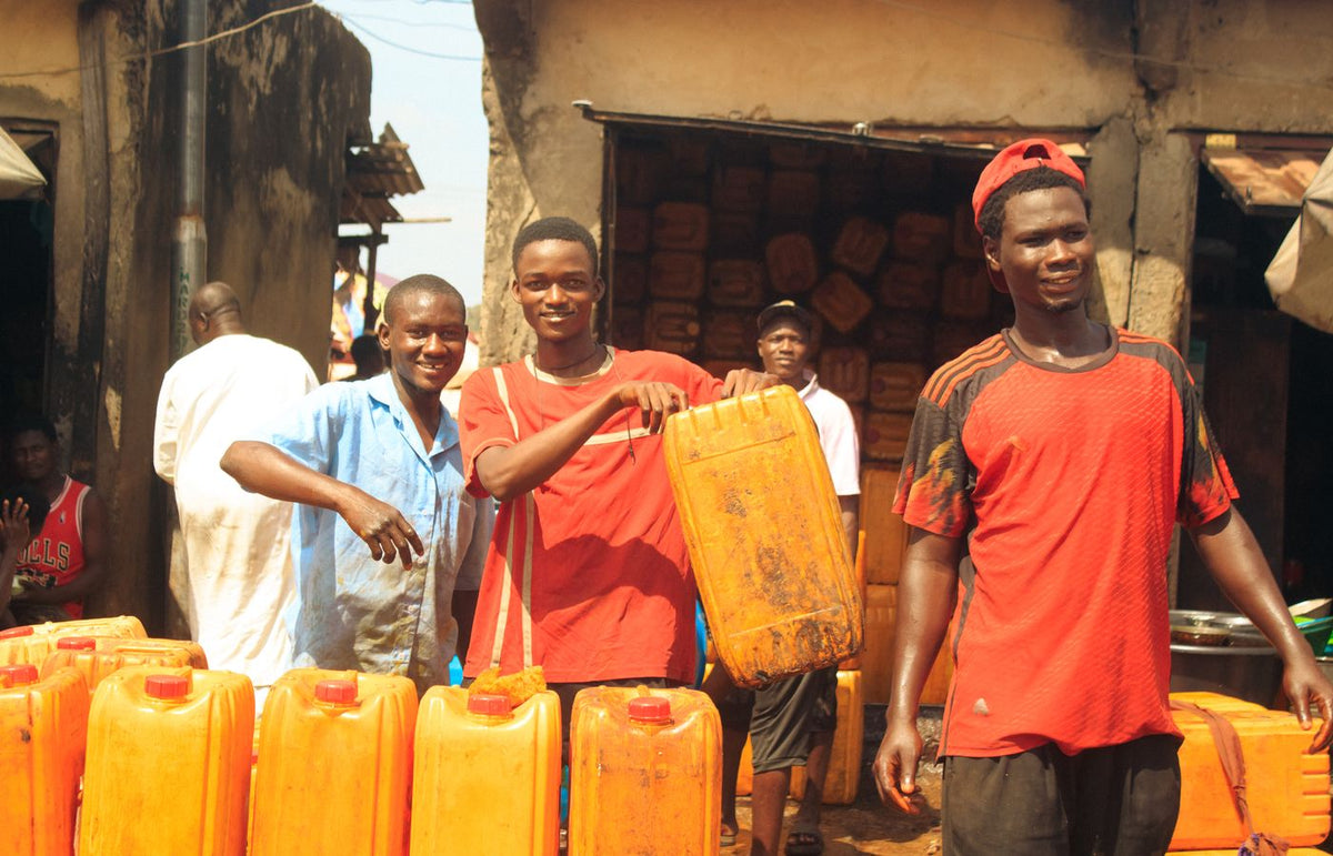 Three young men smiling while handling water containers in a vibrant outdoor market setting.