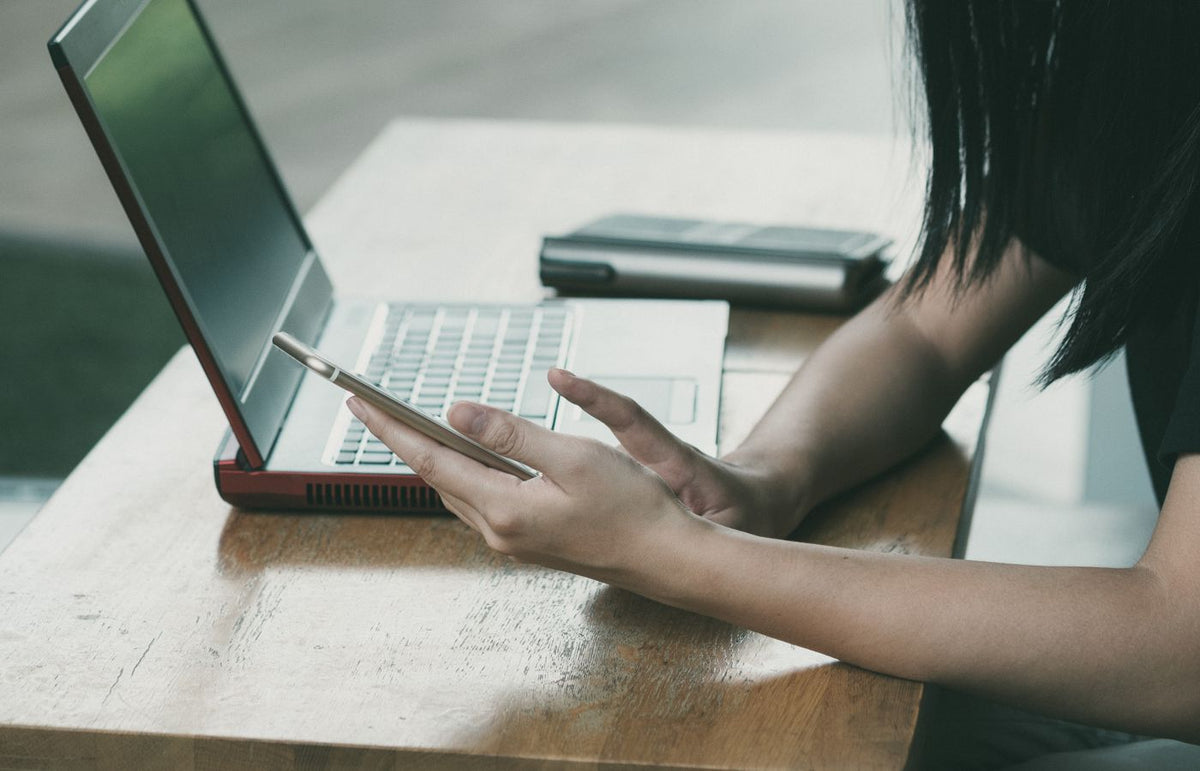 A young woman multitasking with her smartphone and laptop at a table indoors.