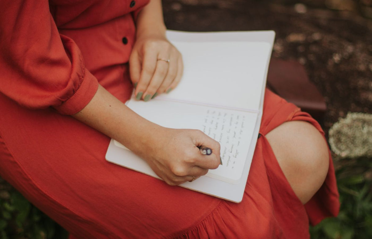 A woman in a red dress writing in a journal, outdoors over a leafy background.