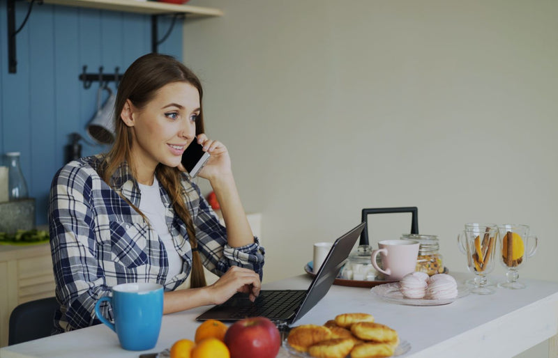 Young woman multitasking on phone and laptop in a cozy kitchen setting.