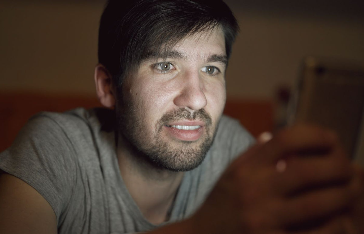 A young adult male engages with a smartphone, illuminated by its screen in a dim setting.