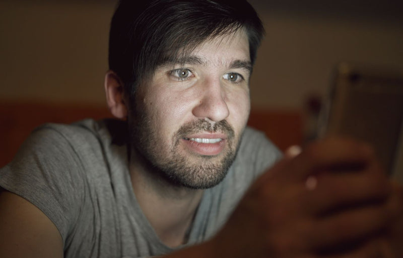 A young adult male engages with a smartphone, illuminated by its screen in a dim setting.