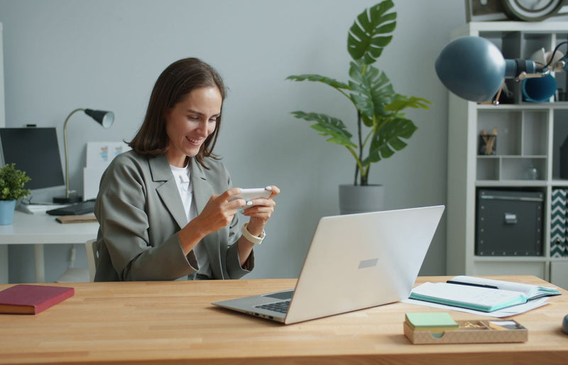 A woman in a home office using a smartphone while working on a laptop.