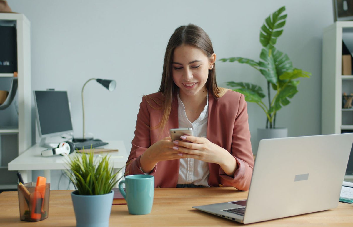Smiling businesswoman checking phone, seated at desk with laptop and plants.