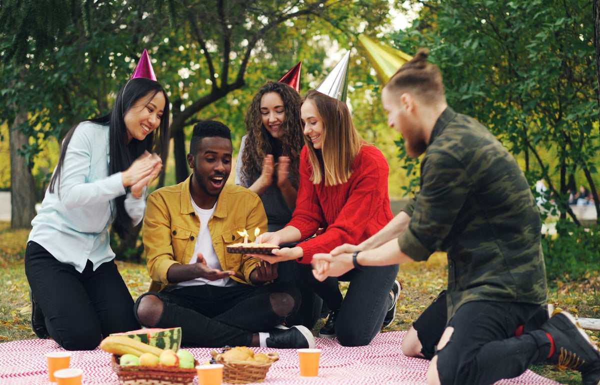 Group of friends enjoying a birthday celebration outdoors with cake and party hats.