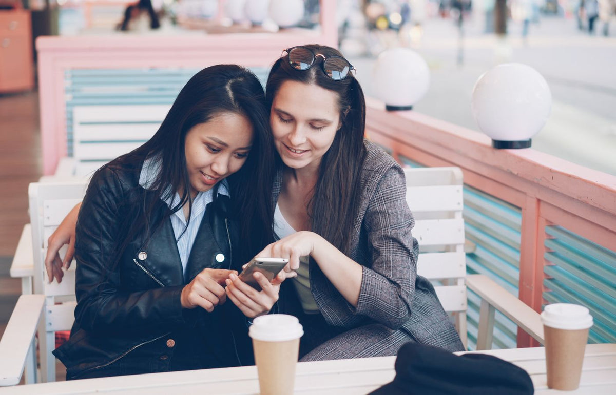 Two women sitting at an outdoor café, sharing a moment with coffee and a smartphone.