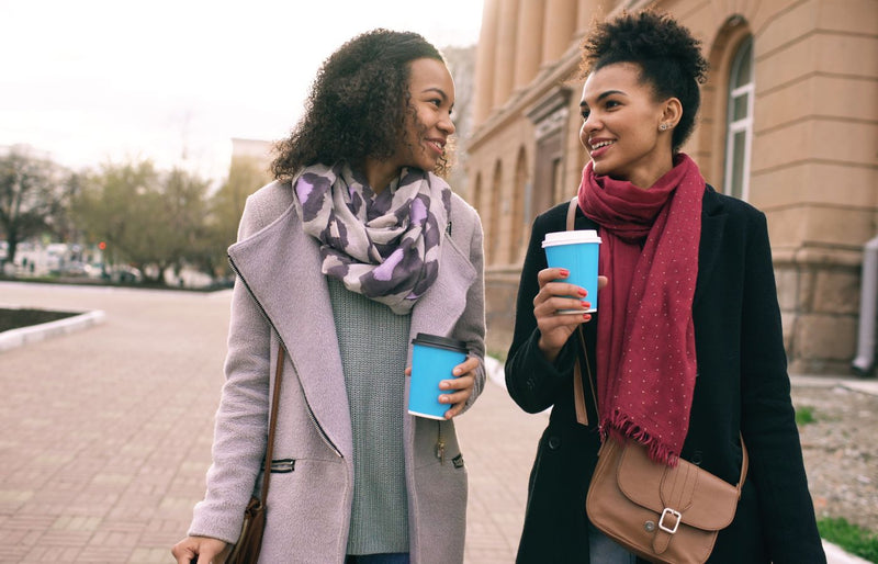Two women walking outdoors with coffee cups, enjoying a conversation.