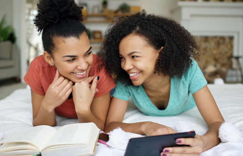 Two young women enjoying reading and using a tablet together indoors on a cozy day.