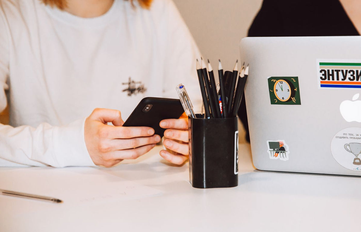 Young adult at a desk texting on smartphone, next to a laptop and stationery holder.