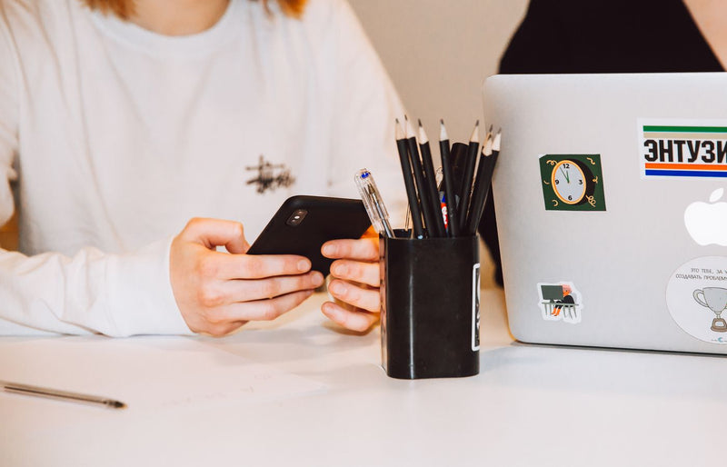 Young adult at a desk texting on smartphone, next to a laptop and stationery holder.