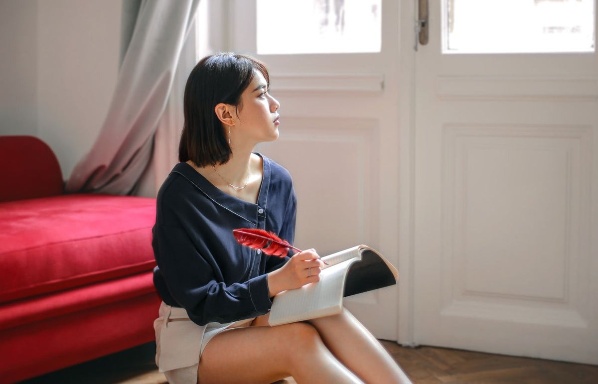 A young woman sits thoughtfully by a window, holding a feather pen and notebook, in a serene room.