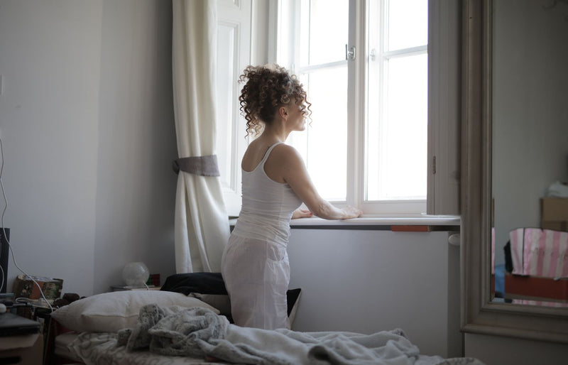 A woman in a bedroom gazes out the window, creating a calm and serene atmosphere.