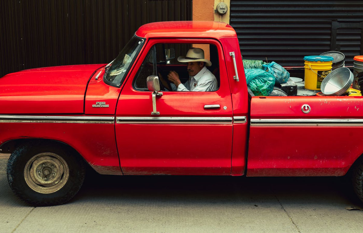 A classic red truck with a driver wearing a hat, parked in an urban area with colorful containers.