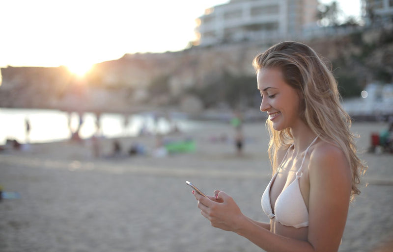 A young woman in a bikini smiles while texting on a sunny beach during sunset.