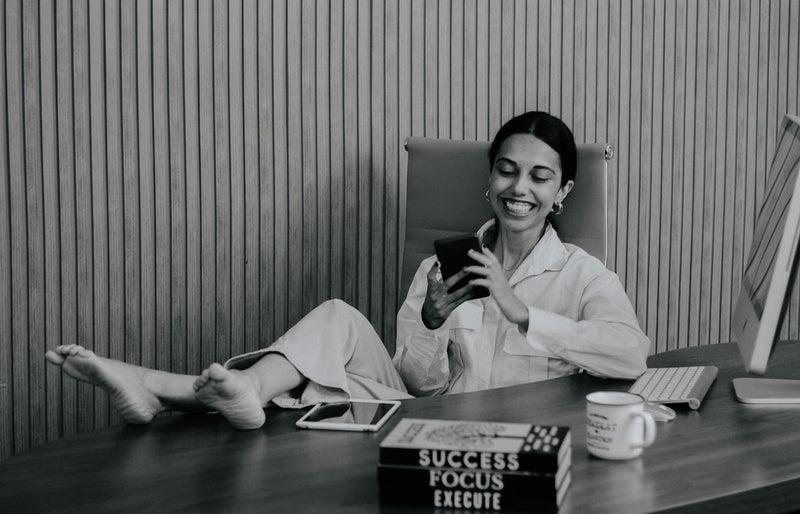 A woman relaxing at her desk with feet up, smiling at her phone.