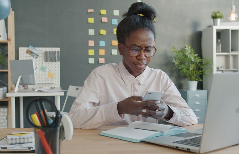 Young woman using a smartphone at her desk, surrounded by office supplies and a laptop.