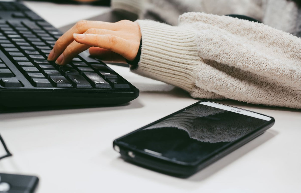 Close-up of a woman's hand typing on a keyboard with a smartphone placed on the desk.