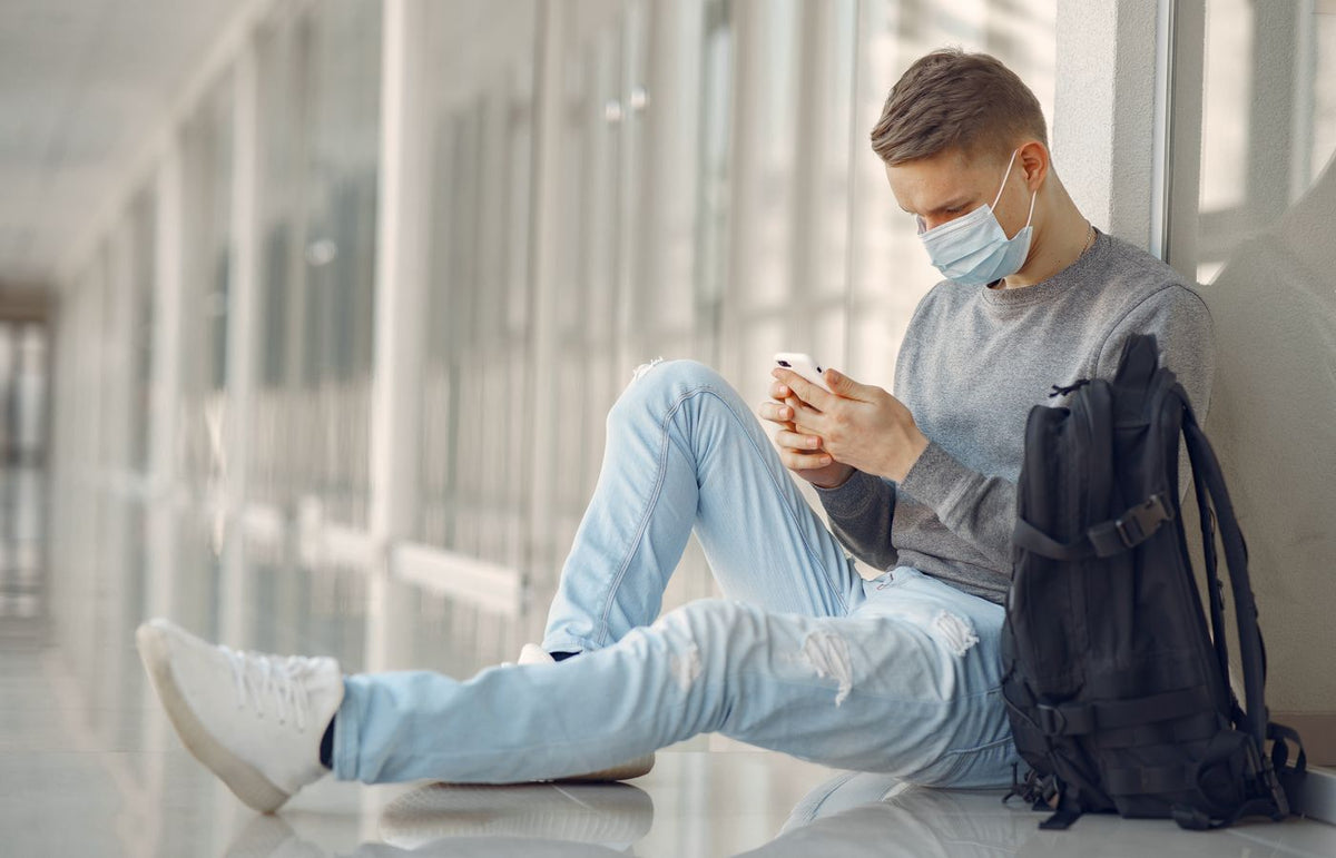 Man in a mask sitting in a hallway, using smartphone, with backpack nearby.