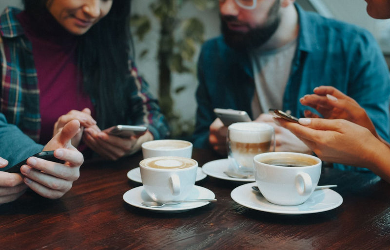 Four adults using smartphones around a coffee table, enjoying various coffees in a cozy café setting.