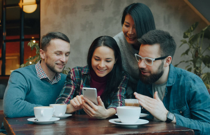 Four friends sharing a moment over coffee in a cozy cafe setting, laughing and engaging with a smartphone.