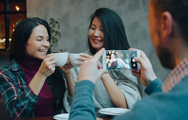 Two friends sharing a joyful moment over coffee indoors with one taking a photo.