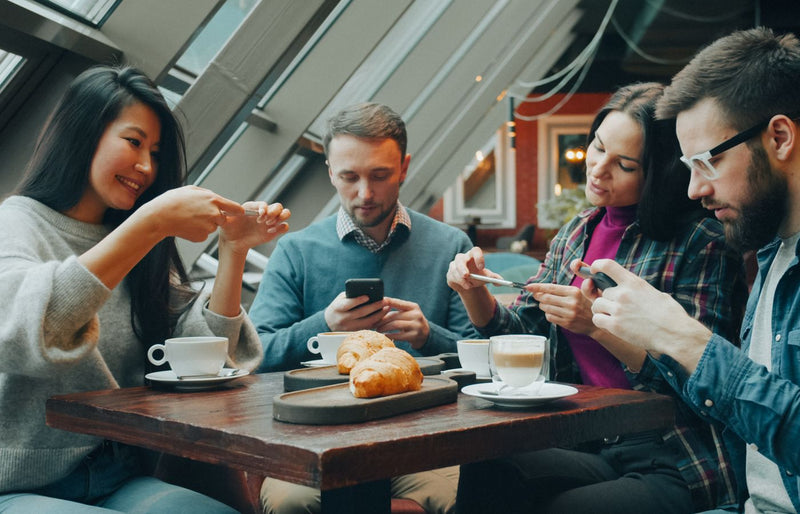 Group of friends interacting with smartphones while enjoying coffee and pastries in a modern café.