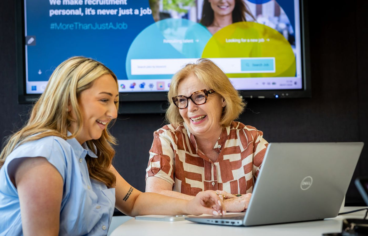 Two women laughing while discussing work at a desk with a laptop in an office.