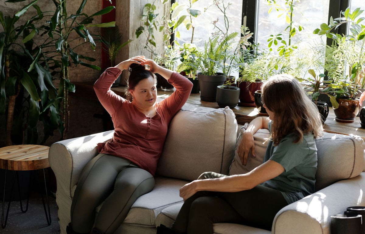 Two women enjoying a relaxed conversation on a sofa in a bright room filled with plants.